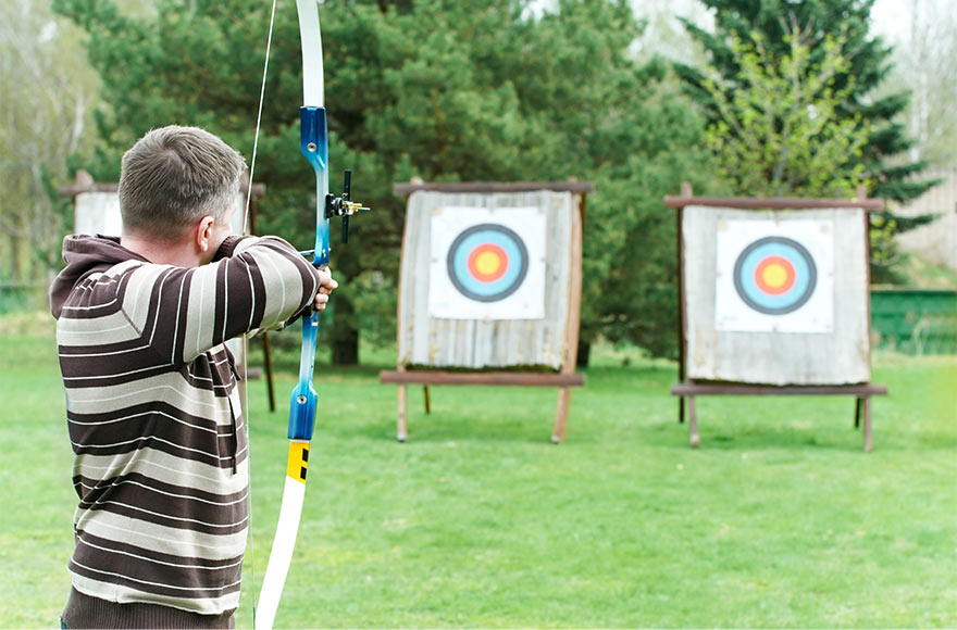 Man in striped hoodie aiming bow and arrow at a target in green open space