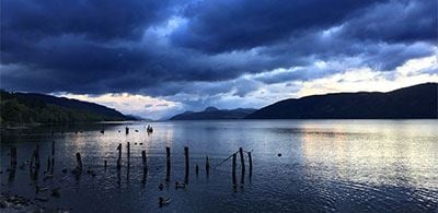 The calm waters of Lock Ness under a dark cloudy sky surrounded by silhouetted hills 