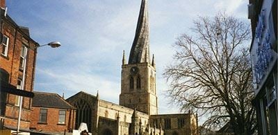 The twisted spire of Crooked Spire Church surrounded by town builidings and a cloudy blue sky 