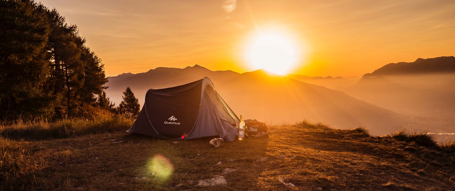 Sunrise over distant mountains with tent in the foreground next to a collection of pine trees 