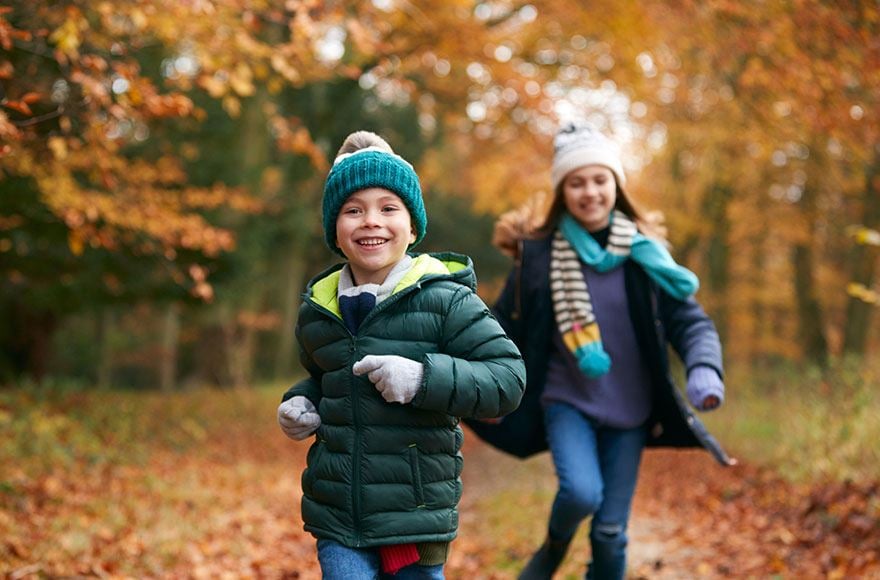 2 young children run down a woodland path covered with fallen autumnal leaves 