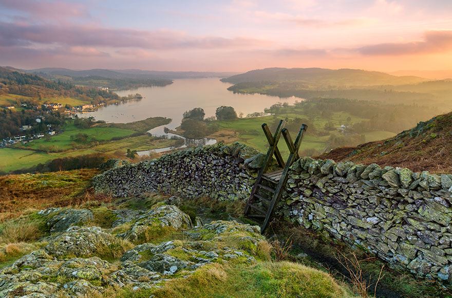 a walking sty placed over a stone walk infront of vast green fields and a calm lake of water 