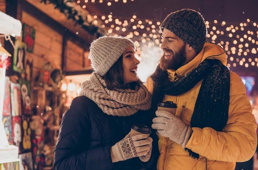 A couple wrapped up in coats and hats gaze lovingly at each other as they hold hot drinks at a Christmas market 