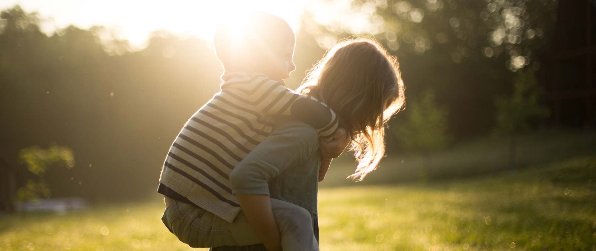 Toddler giving another child a piggyback in sunny green open space surrounded by trees