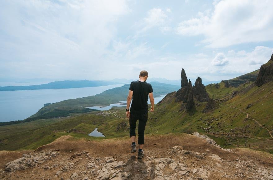 A man walks down a rocky hill aginst the impressive green hills and calm sea