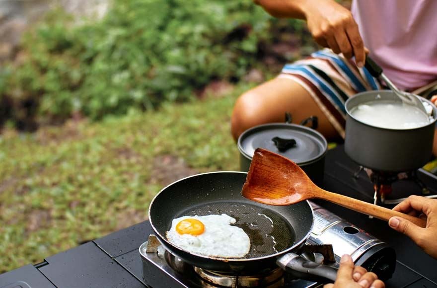 A man sitting down frying an egg in a frying pan over a camp stove