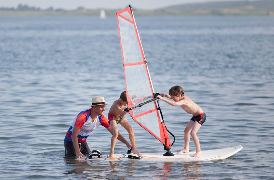 A man helping two children balance on a winder surfing board in a calm lake 