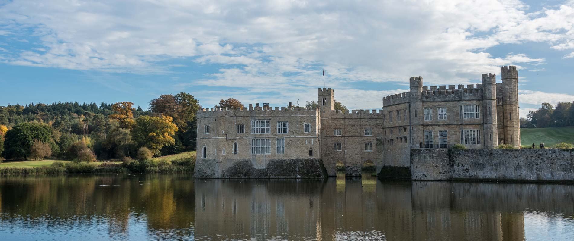 Leeds castle surronded by a lake with a forest of green and yellow autumnal trees nestled behind it