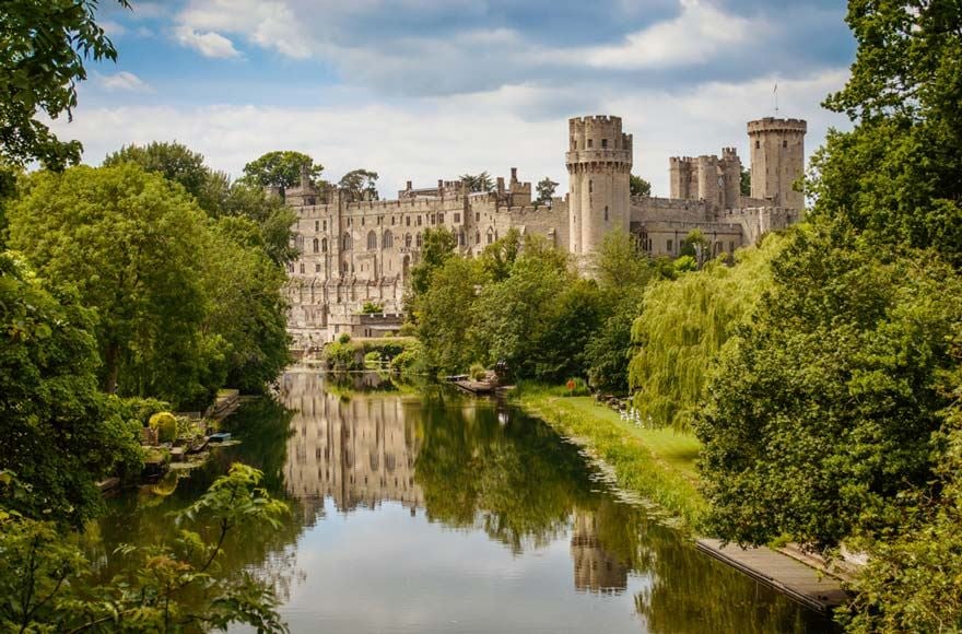 Warwick castle surrounded by a forest of green trees lining a calm lake 