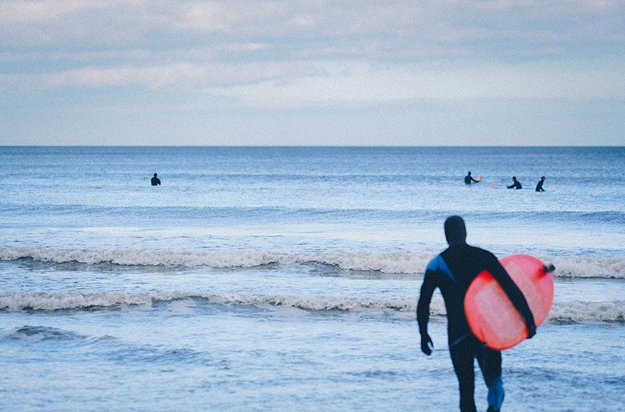 a surfer in w wetsuit holds a red surf board as they walk into the sea with 3 other surfers further out to sea 