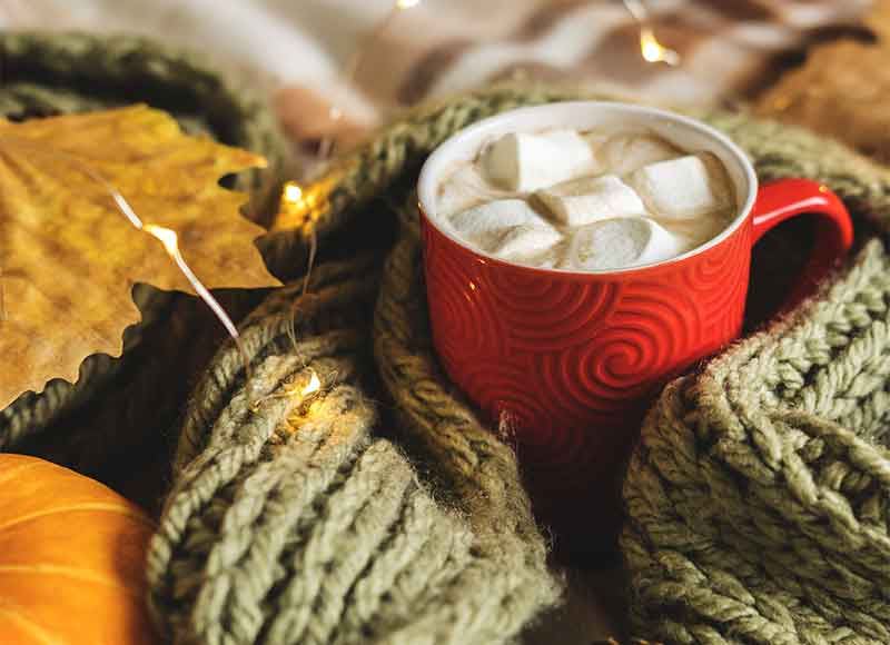 Close up of a mug filled with hot chocolate and marshmallows, surrounded by cosy blankets