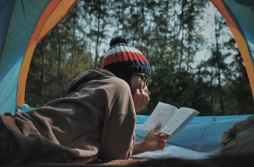 a woman relaxing and reading a book  sitting in a tent with a backdrop of forest trees 