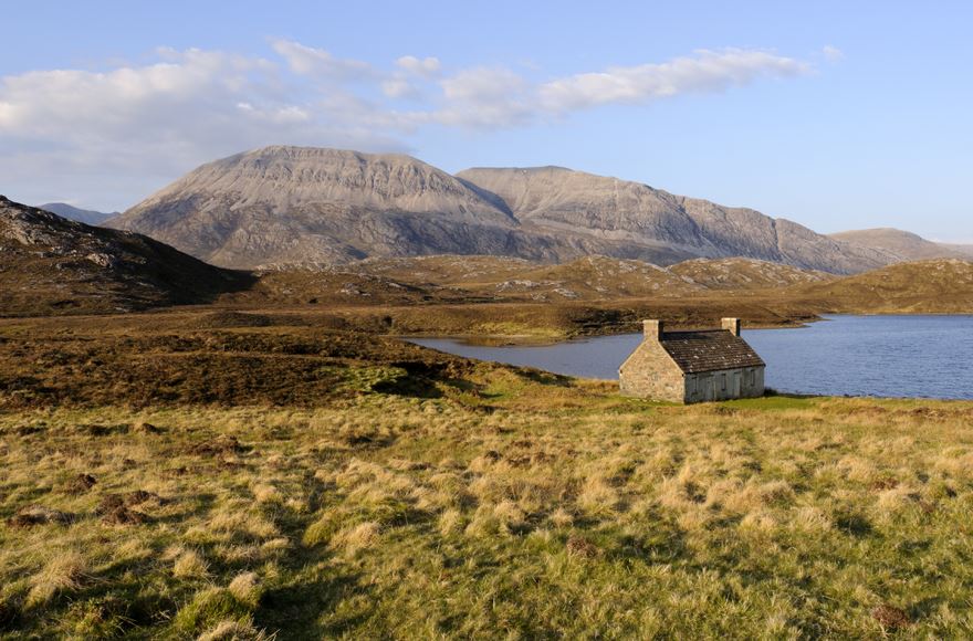 Natural tones of greens and browns from the rugged grasses with a small stone cottage to the right just in front of Loch Shin with towering mountains in the distance, pale blue sky with some white clouds at Loch Shin Sutherland Highlands