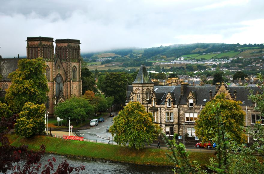 Partial view of a rive in the forefront with Inverness Cathedral across the rive to the left. Stone buildings to the right with a road between the two. Houses then fields  in the background