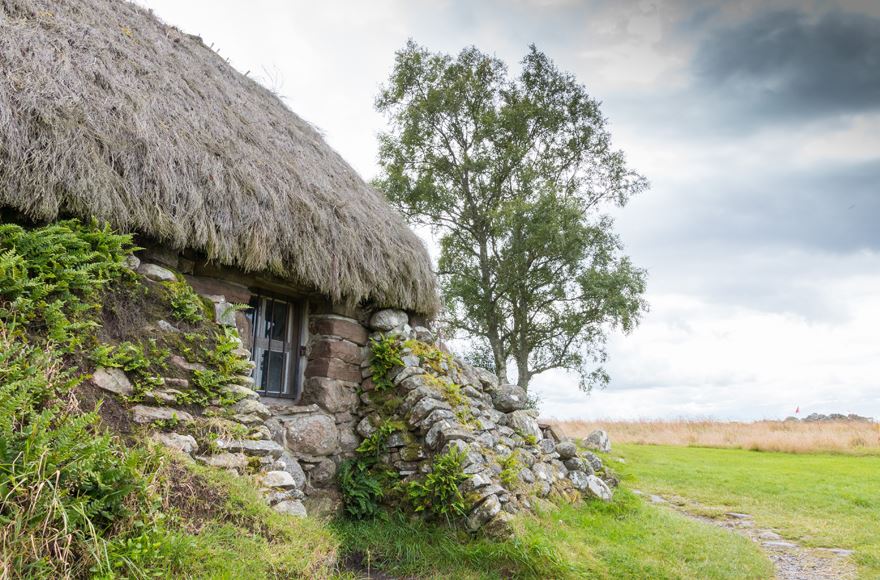 Partial stone cottage to the left with a thatched roof awith two slanted stone walls leaning again it, green grass and a tall tree in the background with greyish sky