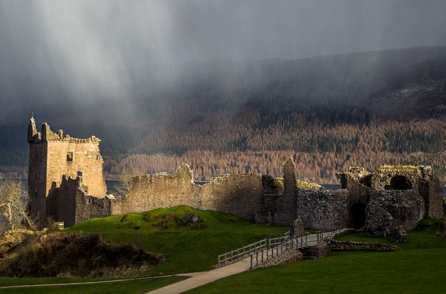 Grean grass with a small bridge leading to the right, with the ruins of Urquhart castle to the left with the sun beams hitting the castle making the brownish trees in the background look a little haunting. 