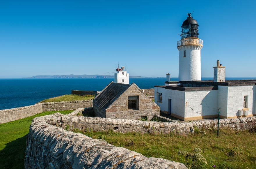 Stone wall with rought grass in the inner field, Over the far side a small stone cottage next to a white swaure building with a lighthouse, with the sea behind it leading to an island in the far distance