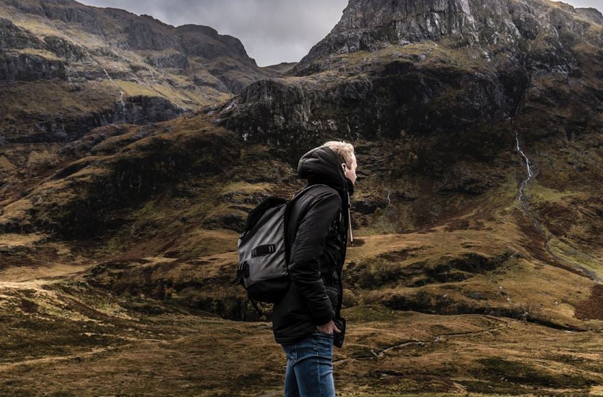 In the foreground in the middle a walker in a black jacket and jeans facing the right  looking up to the close up mountains with brown green grass leading up the rocky faces towering up behind in the Scottish Highlands