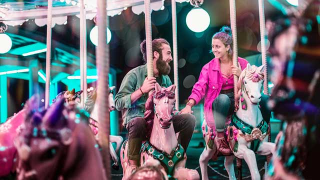 A man with a beard and a woman in a pink shirt ride horses on a carousel