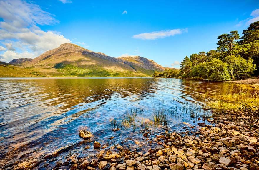 Pebbles in the foreground leading to rippling water of the loch trees to the right, mountains in the background with blue sky