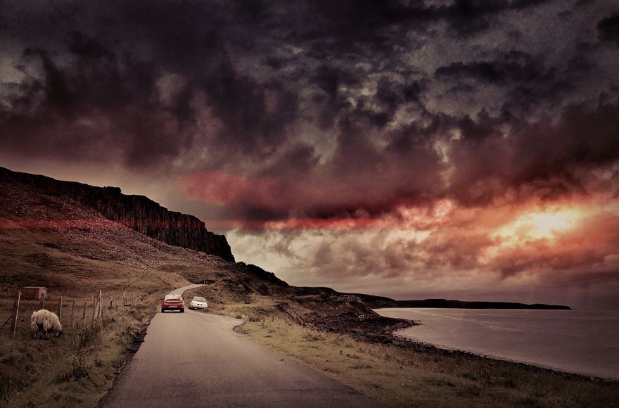 Narrow road by the loch with two cars passing with an amazing sky that looks like a painting. Sun going down  with grey clouds leading to reddish then dark charcoal clouds