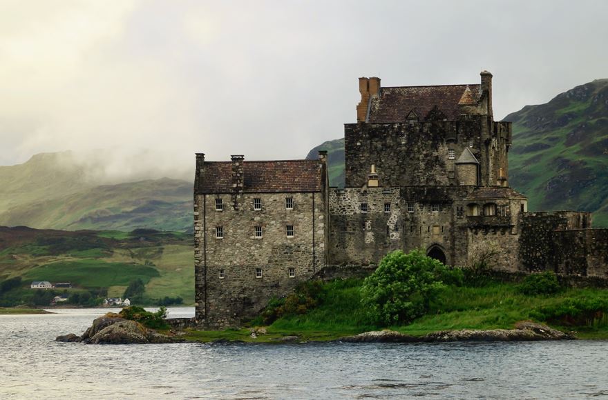 Slight choppy water in the foreground with a dark stone castle, Eilean Donan castle with green hills and misty clouds behind