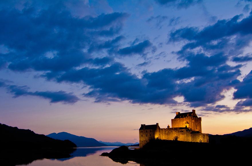 Lights reflecting off the castle while the sun sets in the pale sky which makes the clouds look blue with the silhouette of the mountains along the calm water