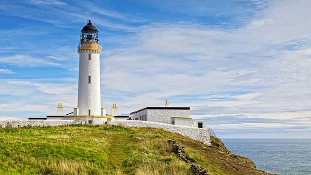 The white Mull of Galloway lighthouse on top of a green hill  reachng up to the blue cloudy sky with the sea to the side