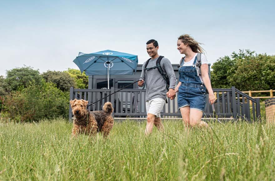 Young couple walking their dog in a lush green landscape with a glamping pod in the background