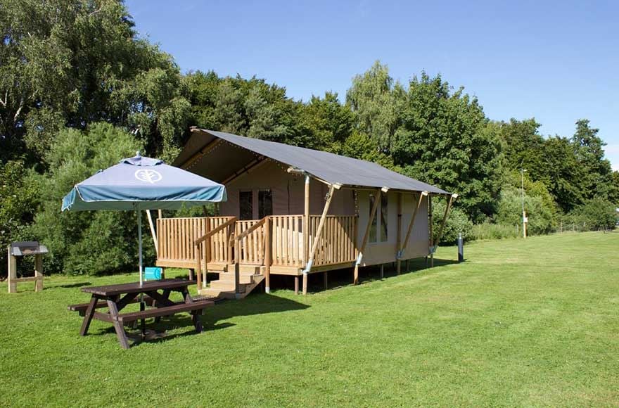 Safari tent and picnic table set up in a grassy field, surrounded by trees and a clear blue sky