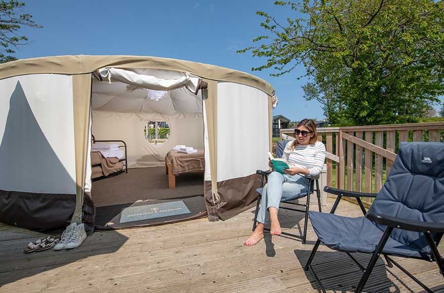 A lady relaxing in a chair outside a canvas yurt, surrounded by trees.