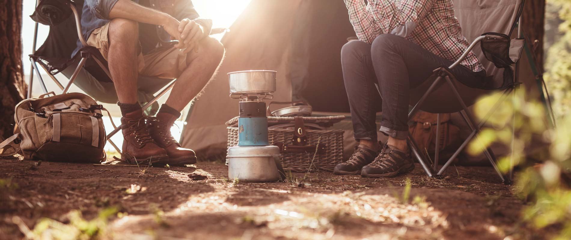 Two campers sit outside a tent, preparing a meal on a camping stove, enjoying the comfort of their outdoor setup