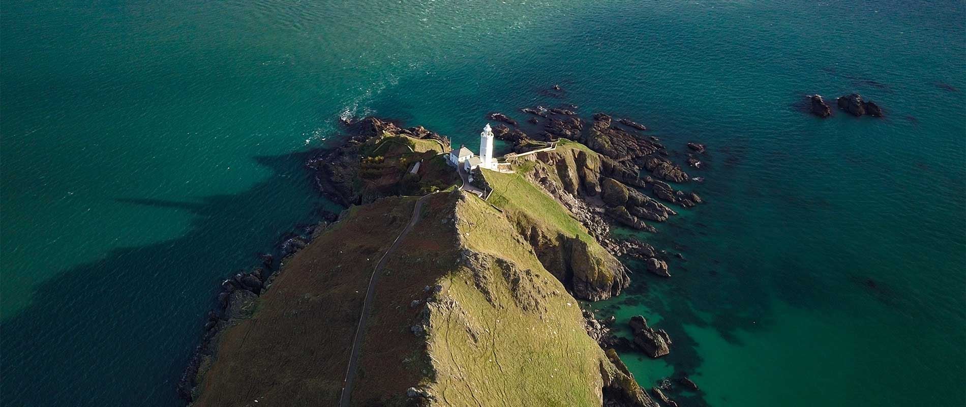 aerial view of the white building of start point light house on the middle of  a rocky island surrounded by dark blue sea 