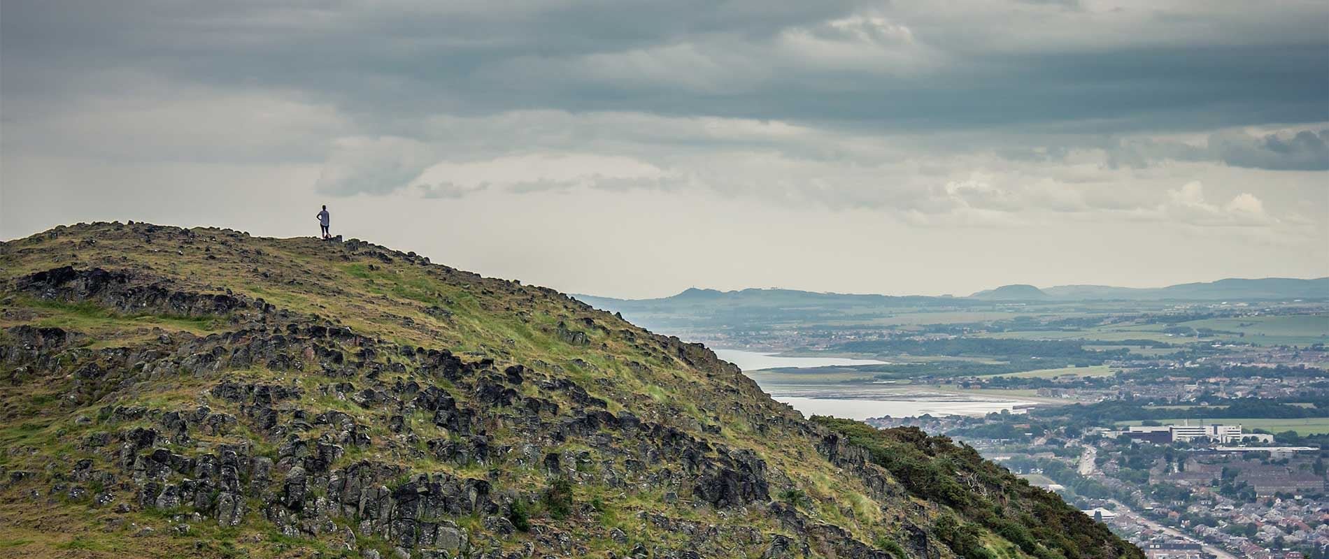 a person stand top the rocky hill of Arthurs seat looking over the city of Edinburgh 