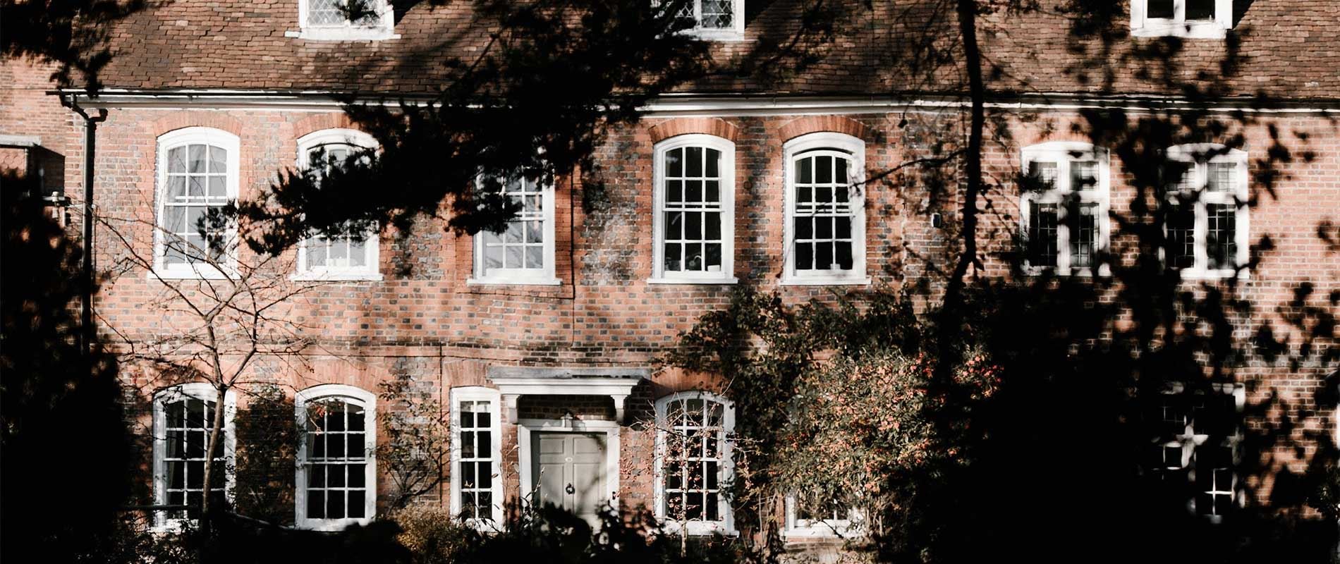 historic brick house with large white windows, framed by shady trees 