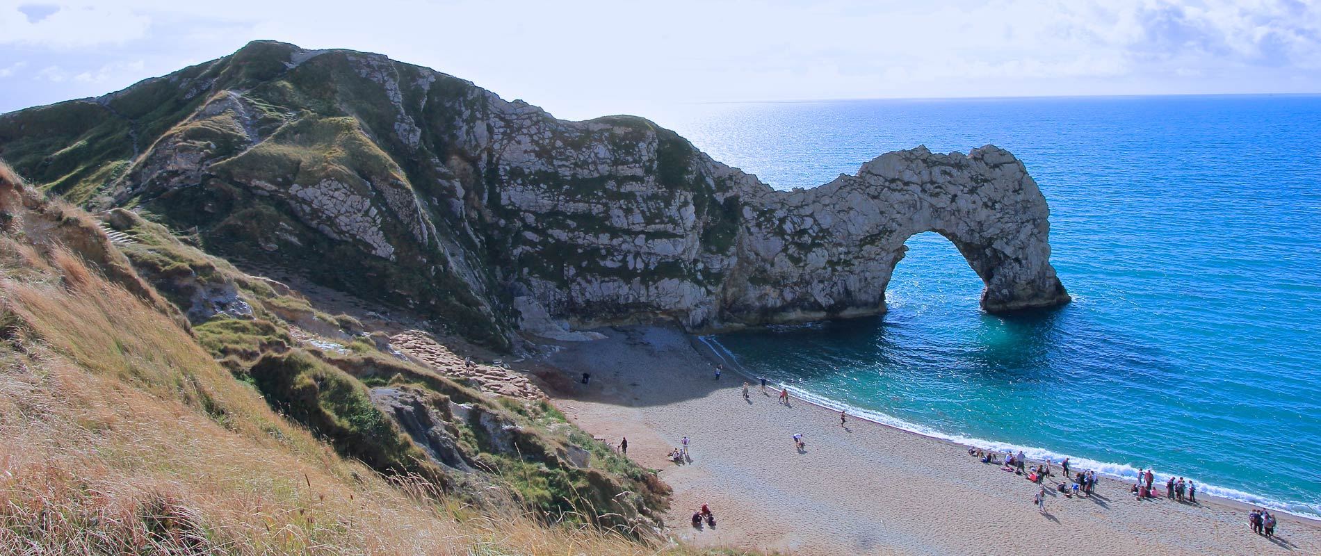 iconic rocks of durdle door in a vibrant blue sea with people walking along the sandy beach 