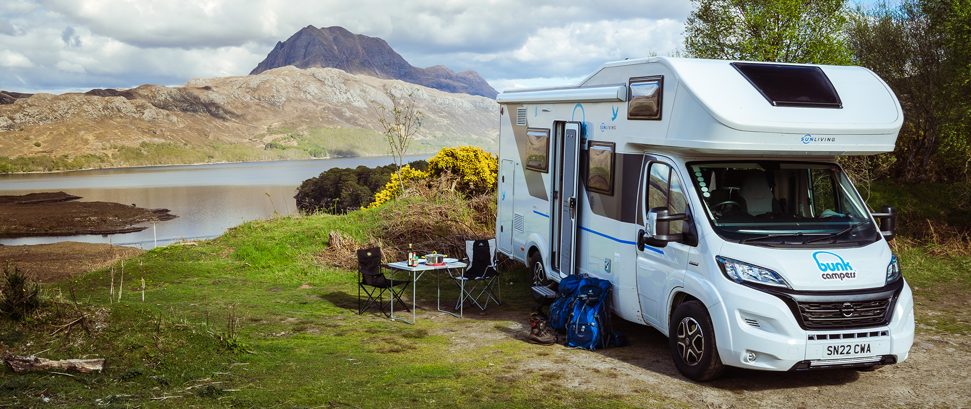 Cmaping table and chairs set up outside a motorhome with a lake and hills in the background