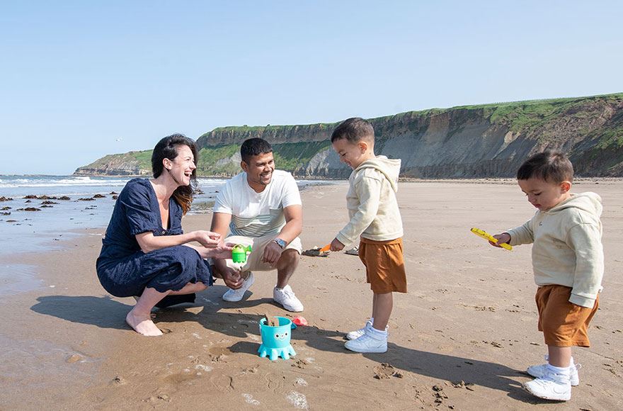 a mother and father crouch down on a sandy beach while their two young sons play in the sand 