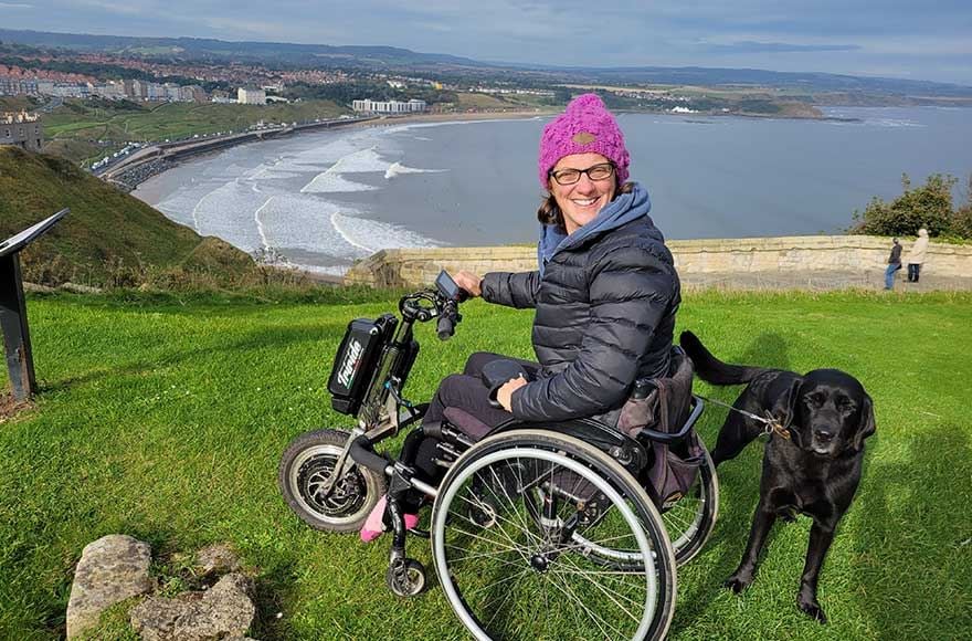 A woman in a wheelchair sits on a hilltop, with her dog beside her, enjoying the beautiful landscape