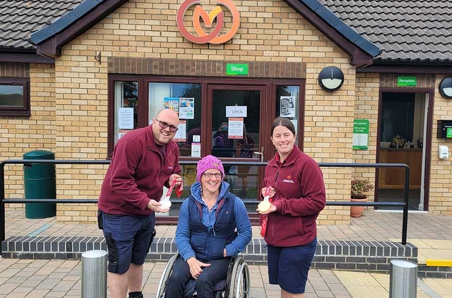 a woman in a wheelchair poses for a photo with 2 members of site staff outside the reception building