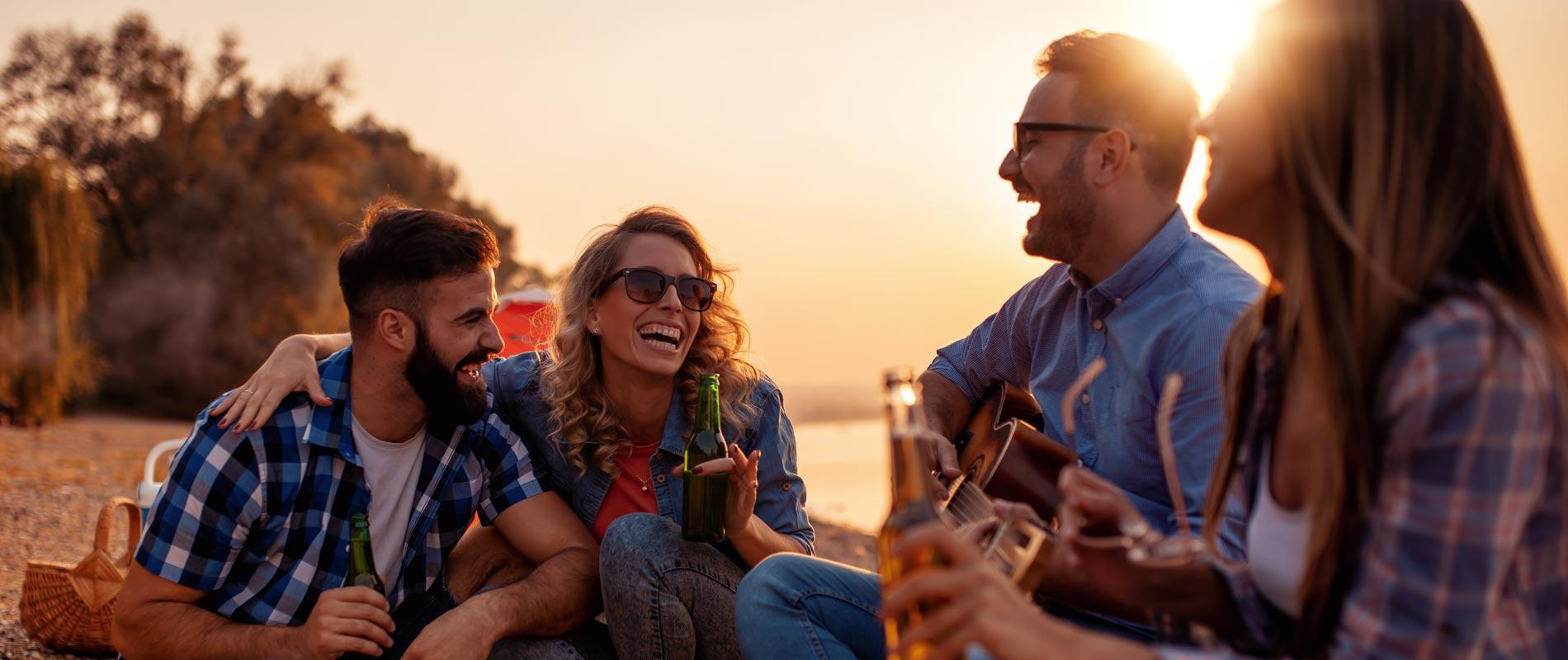 Four friends laughing with beers and guitar playing at campsite