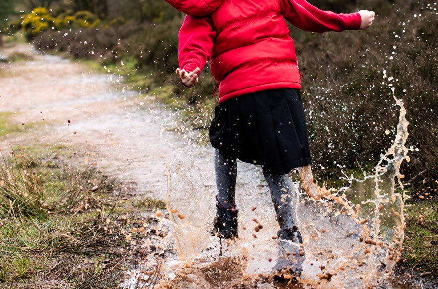Young girl splashing around in puddles on a muddy path, wearing wellington boots