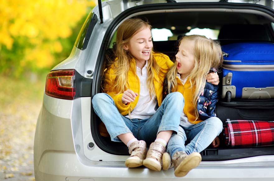 Two girls sitting on edge of car boot as family pack